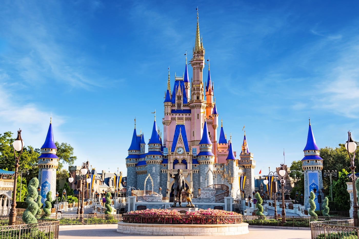 Castelo da Cinderela no Magic Kingdom em dia de céu azul, com jardins floridos e a estátua de Walt Disney e Mickey